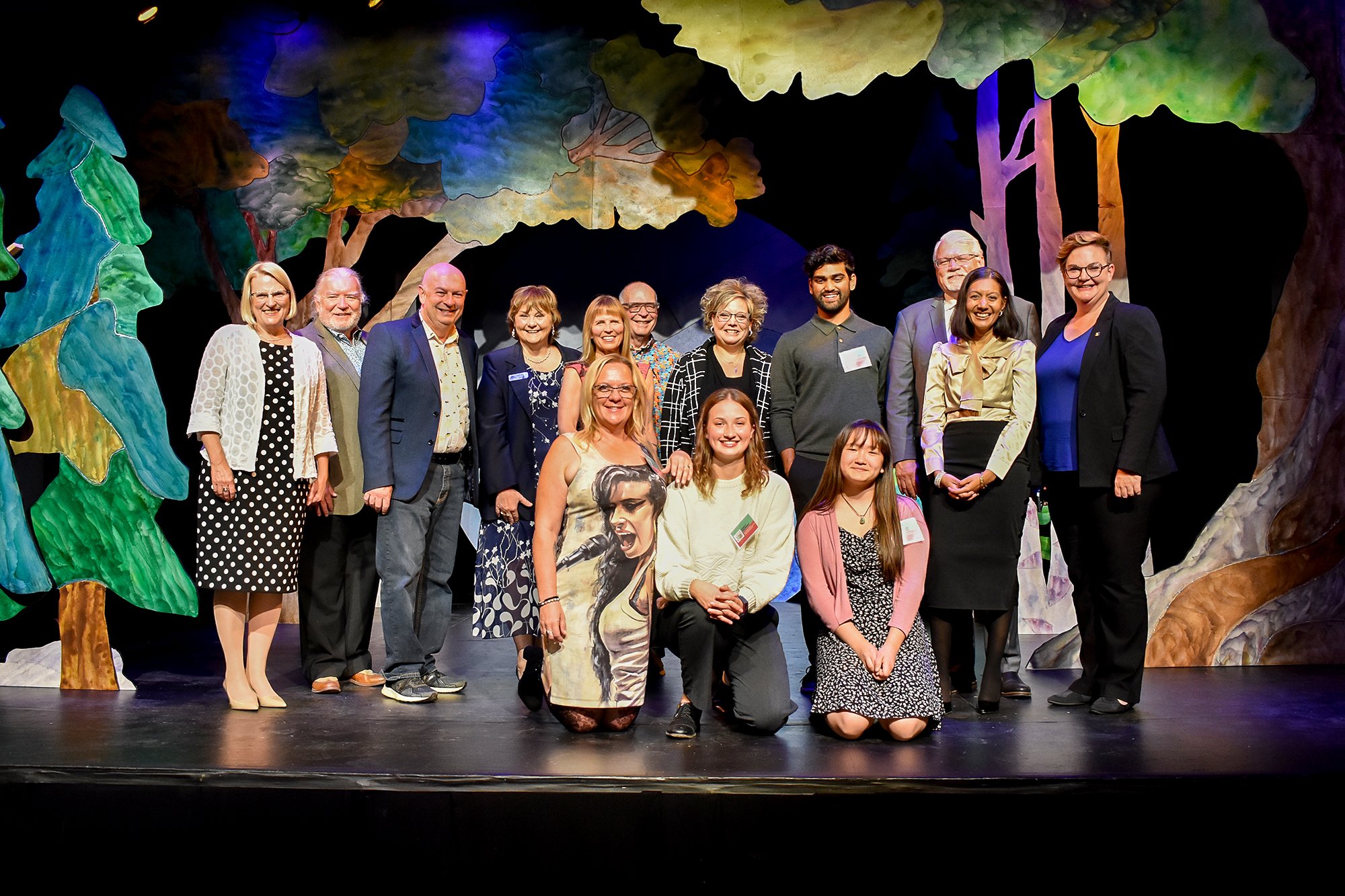 A group of presenters and award winners stand on a stage at Theatre Orangeville. They are surrounded by a beautiful set of tall, watercolour designed trees.